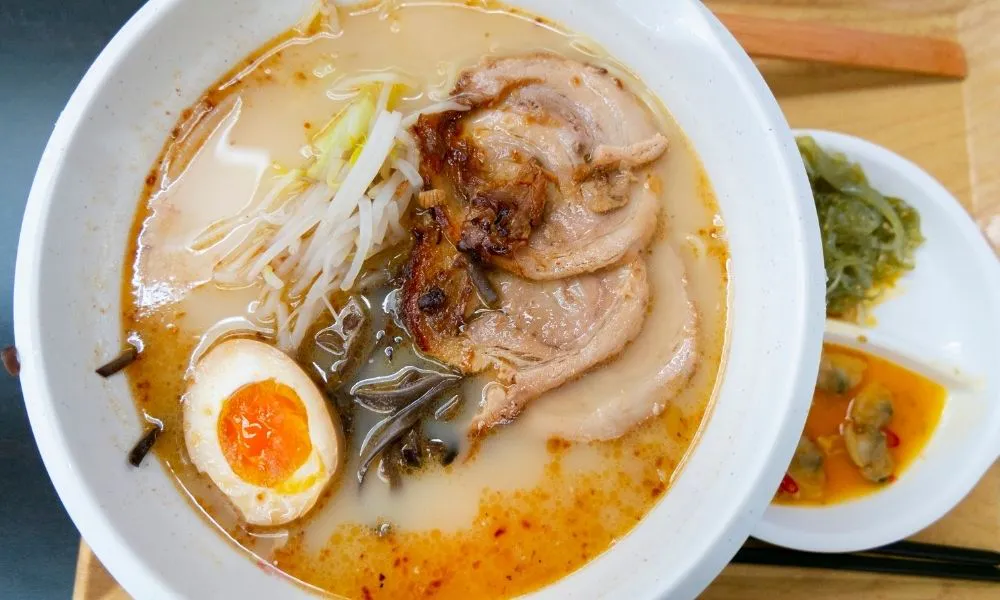 Close‑up of tonkotsu ramen in white bowl, creamy pork broth with chashu pork slices, soft‑boiled ajitama egg, bean sprouts and wood ear mushrooms, side dish of seaweed salad and spicy clams on wooden table.