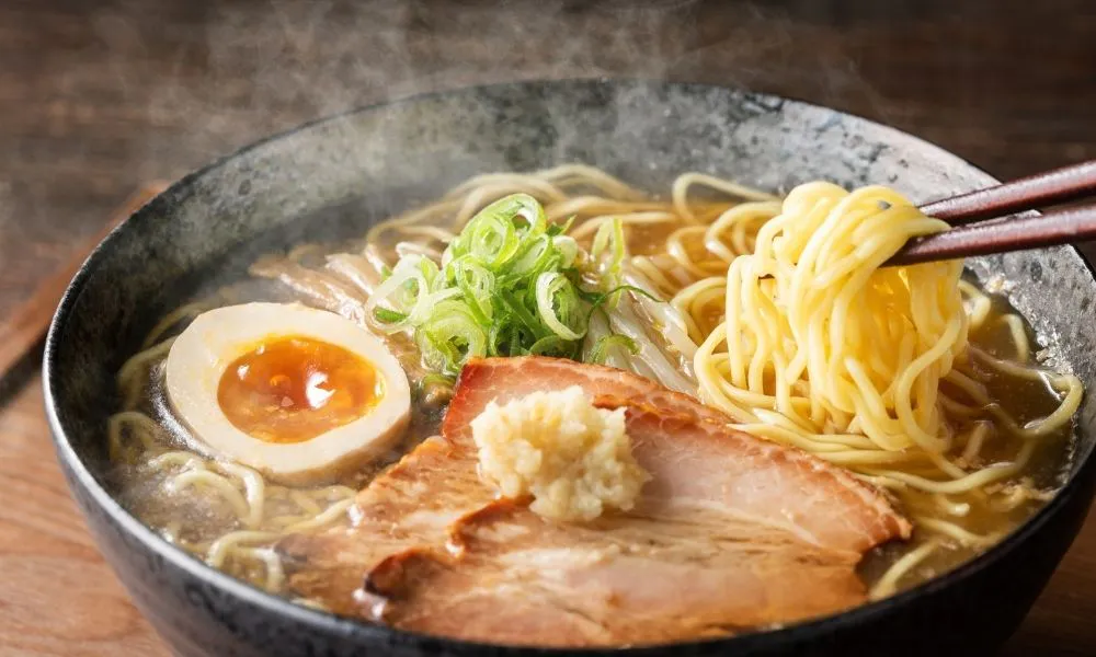 Steaming shoyu ramen in black ceramic bowl, brown soy broth with curly noodles, thick chashu pork slice topped with grated daikon, halved soft‑boiled egg, chopped green onions, chopsticks lifting noodles.