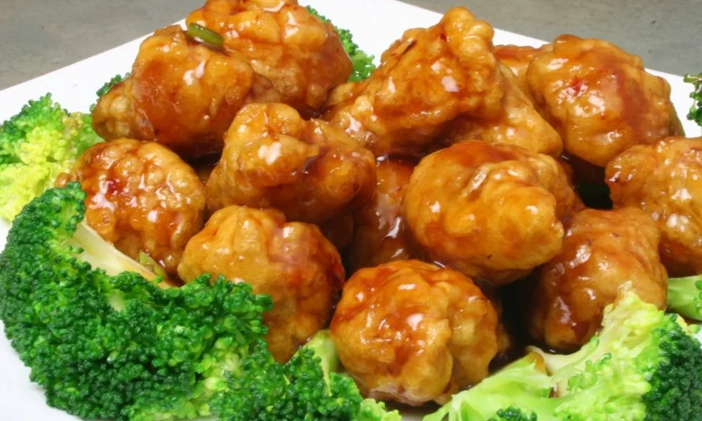 General Tso's chicken, golden fried chicken chunks coated in glossy reddish‑brown sweet‑savory sauce, arranged on white plate surrounded by fresh green broccoli florets, close‑up food shot.
