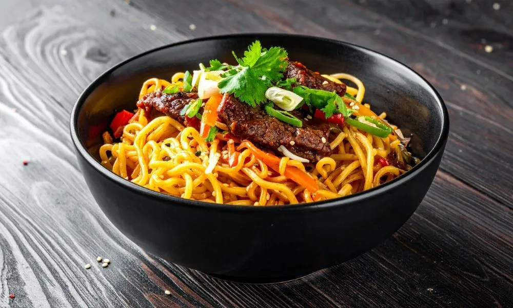 Beef lo mein stir‑fried noodles in black bowl, thick yellow noodles tossed with sliced beef, red chili, carrot strips, green onion and fresh cilantro, sesame seeds sprinkled, dark wooden table background.