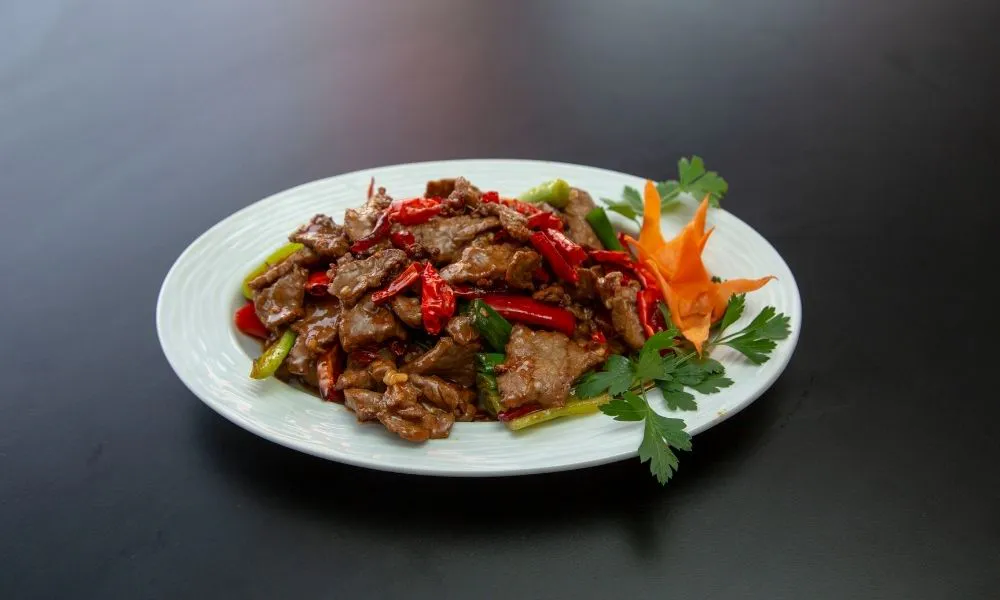 Spicy Hunan beef stir‑fry on white oval plate, tender beef slices with dried red chili peppers, green celery strips, carved carrot flower garnish and parsley leaves, dark smooth tabletop.