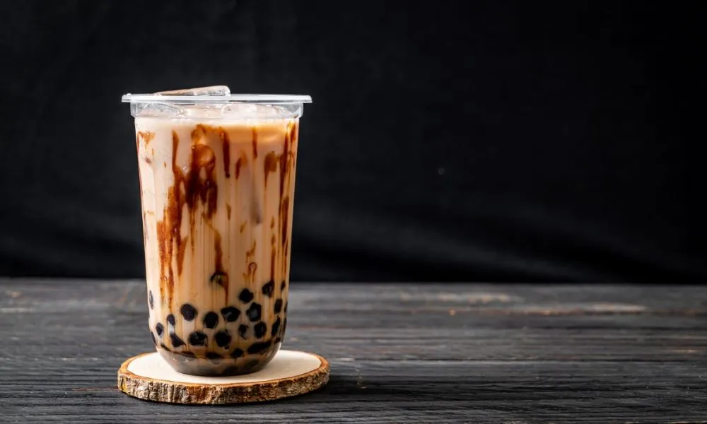 Brown sugar boba milk tea in clear plastic cup with lid, dark brown sugar syrup streaks on cup wall, black tapioca pearls at bottom, ice cubes on top, placed on wooden coaster over dark wood table.