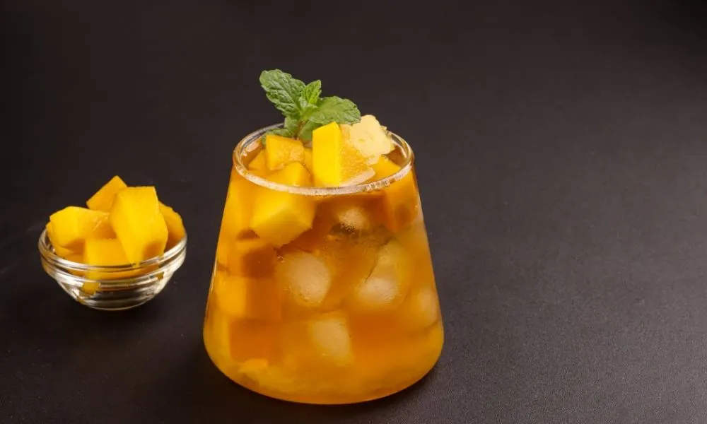 Mango iced tea in short wide glass, golden amber tea with ice cubes and fresh mango chunks, mint sprig garnish on top, small glass bowl of mango cubes beside it, dark matte background.