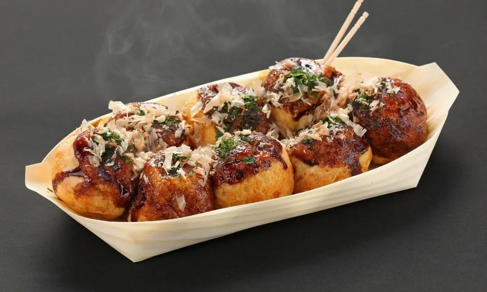 Eight takoyaki octopus balls in wooden boat tray, golden crispy balls drizzled with takoyaki sauce and mayo, topped with bonito flakes and green onion, two wooden skewers inserted, rising steam, dark gray background.